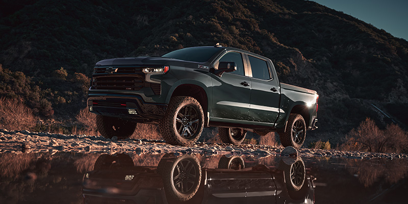 Driver's side view of a green 2025 Chevy Silverado parked in a puddle on rocky terrain with the sun shining overhead
