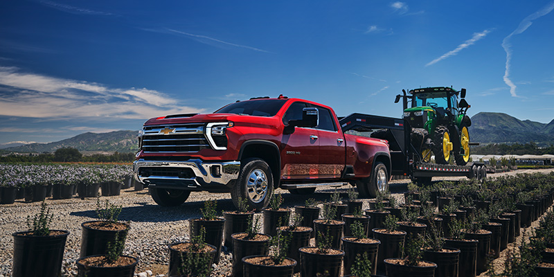 Front driver's side view of a red 2025 Chevy Silverado 2500 HD towing a tractor down a gravel road with large hills in the background and blue skies overhead