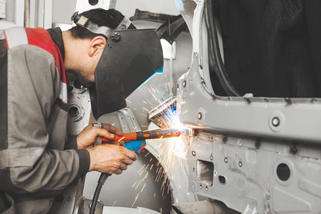 Auto body technician wearing a welding mask repairing a vehicle frame with sparks flying in a workshop