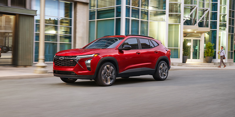 Red 2026 Chevrolet SUV driving on a city street in front of modern glass buildings.