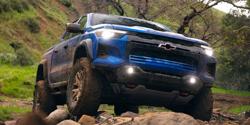 Blue 2026 Chevrolet Colorado driving off-road over rocks with mud on its tires.