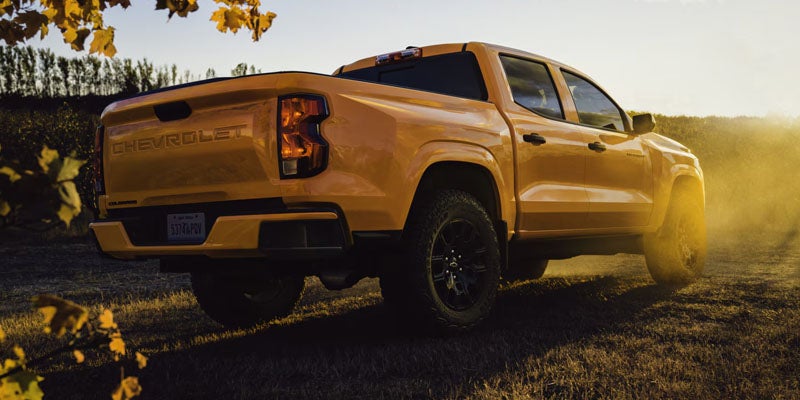 Yellow 2026 Chevrolet Colorado pickup truck driving through a dusty field at sunset.