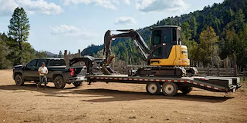 2026 Chevrolet Silverado 2500HD towing a trailer with an excavator in a scenic rural landscape.
